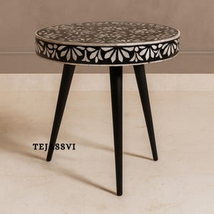 May include: Round black side table with a floral inlaid design on the tabletop. The table has a white border and three black, tapered legs. The brand name "TEJASSVI" is printed on the front.
