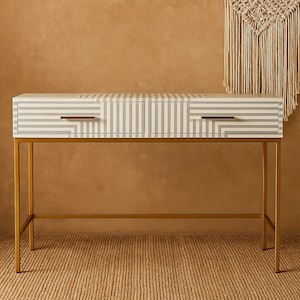 Puede incluir: Un escritorio a rayas blancas y grises con dos cajones y patas de metal color dorado. El escritorio tiene forma rectangular y está sobre una alfombra tejida. Los cajones tienen tiradores dorados.