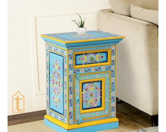 May include: A hand-painted, blue wooden side table with a drawer and door. The table features intricate floral designs in various colors, including yellow, red, and green. The edges are trimmed in yellow, and a small plant sits on top.