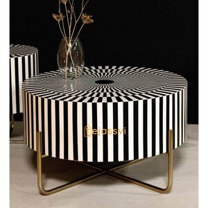 Op de afbeelding: Een ronde salontafel met een zwart-wit streepjespatroon op de zijkanten en bovenkant. De tafel heeft een goudkleurige metalen basis. Een glazen vaas met gedroogde bloemen staat op de tafel. Een modern woondecoratie-item.