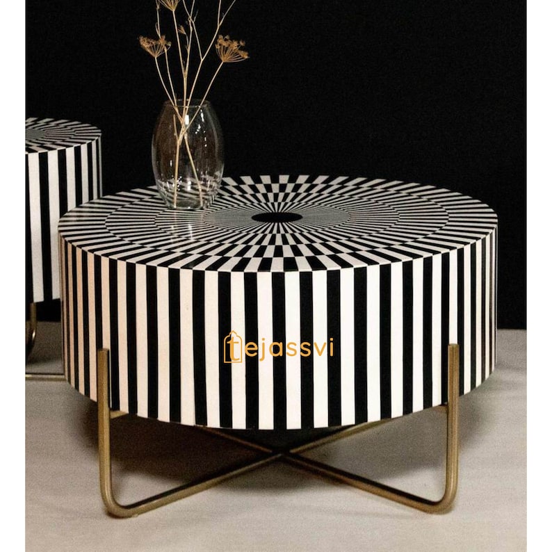 Op de afbeelding: Een ronde salontafel met een zwart-wit streepjespatroon op de zijkanten en bovenkant. De tafel heeft een goudkleurige metalen basis. Een glazen vaas met gedroogde bloemen staat op de tafel. Een modern woondecoratie-item.