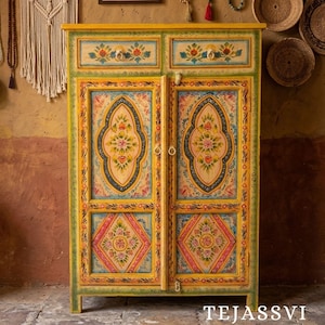 Peut inclure: Un meuble en bois peint à la main avec un cadre jaune et des bordures vertes. Les portes et les tiroirs du meuble sont ornés de motifs floraux roses, bleus et jaunes. Le texte "TEJASSVI" est au bas du meuble.