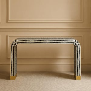 Può includere: Una consolle in bianco e nero dal design curvo. Il tavolo presenta un motivo geometrico ed è impreziosito da dettagli bianchi e dorati. Le gambe hanno una finitura dorata. Questo tavolo decorativo è adatto per ingressi o soggiorni.