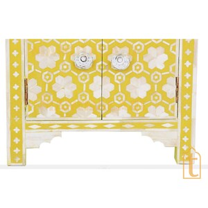 May include: A small, yellow cabinet with a white floral and geometric pattern. The cabinet has two doors with clear, round knobs and decorative white trim. The cabinet is approximately 18 inches wide and 12 inches tall.