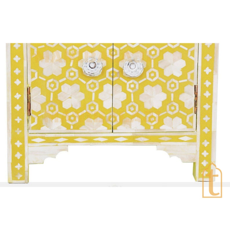 May include: A small, yellow cabinet with a white floral and geometric pattern. The cabinet has two doors with clear, round knobs and decorative white trim. The cabinet is approximately 18 inches wide and 12 inches tall.