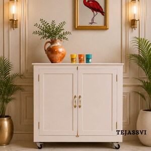 May include: A white cabinet with two doors and brass handles, set against a wall with sconces and a framed bird print. The cabinet has a terracotta vase with greenery and three colorful glasses on top. The text "TEJASSVI" is visible on the cabinet.