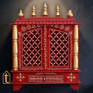 May include: A red wooden cabinet featuring two doors with intricate lattice cutouts, gold-painted pillars on each side, and five gold-painted finials on the top. The base includes a single drawer decorated with gold floral patterns and small white accents.