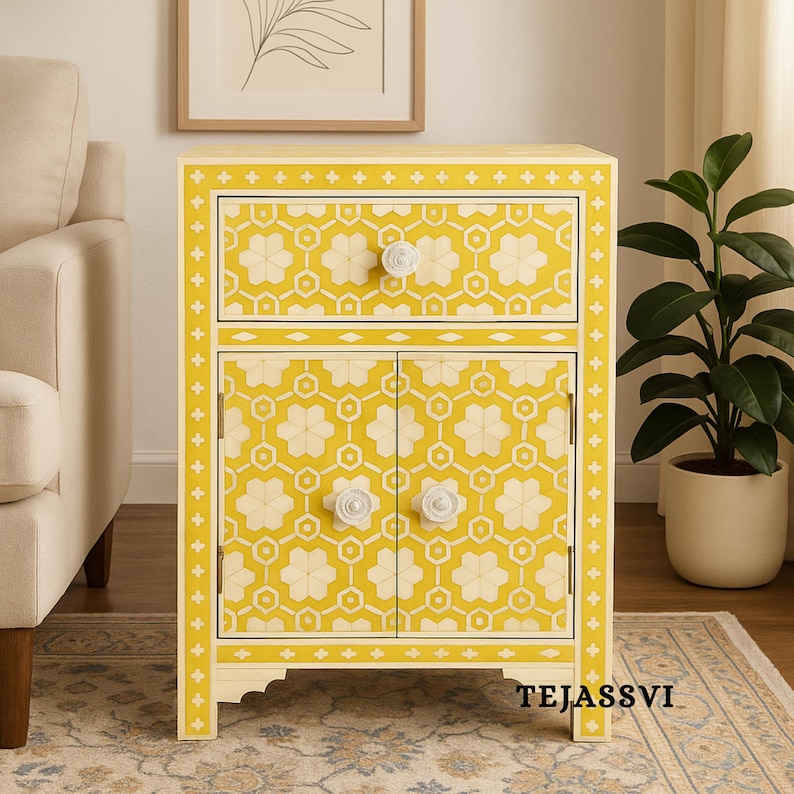 May include: A yellow nightstand with a floral pattern and two doors. The nightstand has a drawer and two doors with white flower-shaped knobs. The floral pattern is cream-colored and the nightstand has a yellow border. The text "TEJASSVI" is at the bottom.