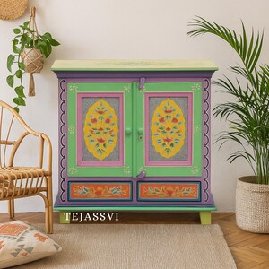 Puede incluir: Un armario pintado con colores vibrantes con cuerpo verde, ribetes morados y diseños florales amarillos. El armario tiene dos puertas y dos cajones, con el texto "TEJASSVI" en la parte inferior. El armario está decorado con detalles rosas, naranjas y amarillos.