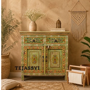 Peut inclure: Un meuble peint à la main avec deux portes et des motifs floraux verts, jaunes et rouges. Le meuble a deux tiroirs et une base vert clair. Le mot "TEJASSVI" est imprimé sur le devant du meuble.