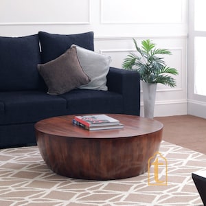 Pode incluir: Uma mesa de centro redonda de madeira castanha escura com uma superfície lisa. Dois livros estão empilhados em cima. A mesa está em frente a um sofá azul marinho com duas almofadas. Uma planta decorativa está no fundo.