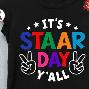 May include: A black t-shirt with the text "It's STAAR Day Y'all" in rainbow colors with white peace signs on either side.
