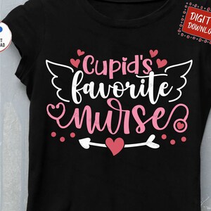 以下が含まれることがあります： 白い文字で「Cupid's favorite nurse」と書かれた黒いTシャツ。ピンクのハートと羽根が付いています。