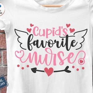 以下が含まれることがあります： ピンクと黒の文字で「Cupid's favorite nurse」と書かれた白いTシャツ。ピンクのハートと羽根が付いています。