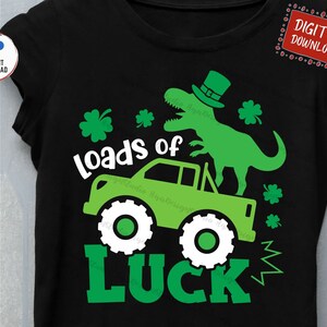 May include: Black t-shirt with a green dinosaur wearing a leprechaun hat, atop a green monster truck. The text "loads of LUCK" is in white and green. Includes a digital download icon.