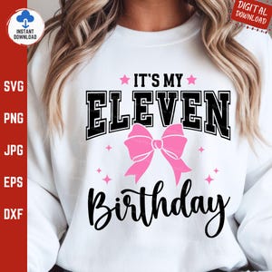 Op de afbeelding: Witte sweatshirt met de tekst "IT'S MY ELEVEN BIRTHDAY" in zwart en roze. Het woord "ELEVEN" staat in een vetgedrukte, collegiale lettertype, met een roze strik eronder. Roze sterren zijn verspreid.