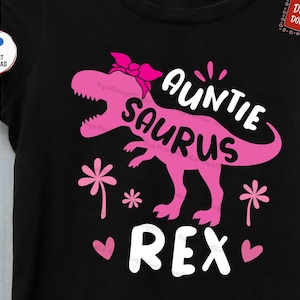 May include: Pink dinosaur silhouette with a bow on its head and the text "Auntie Saurus Rex" in white. The dinosaur is on a black background with pink flowers and hearts.