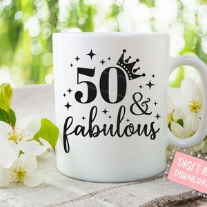 May include: White ceramic mug with the text "50 & fabulous" in black script and a crown design. The number 50 is also in black. The mug has a handle and is set against a blurred green and white floral background.