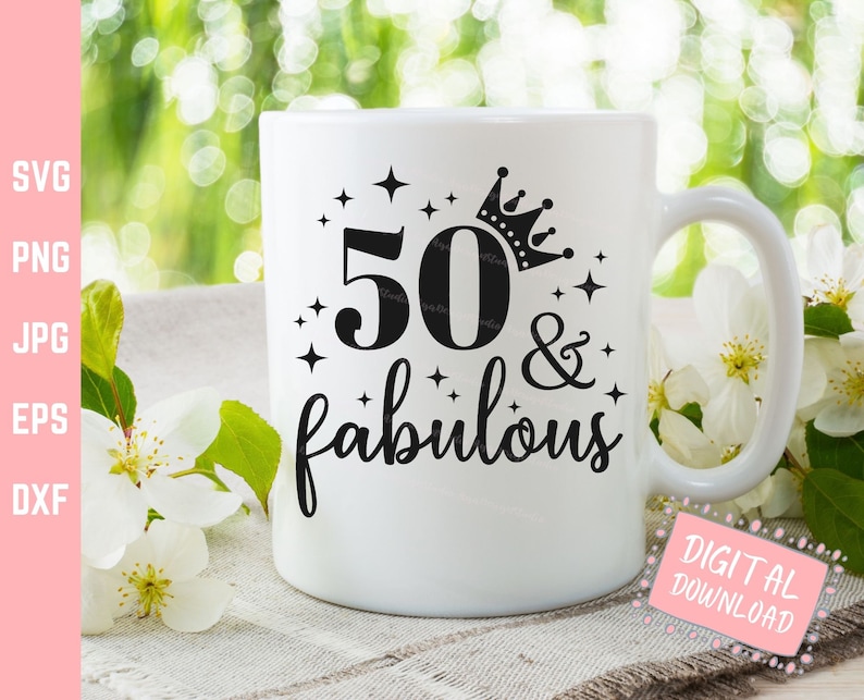 May include: White ceramic mug with the text "50 & fabulous" in black script and a crown design. The number 50 is also in black. The mug has a handle and is set against a blurred green and white floral background.