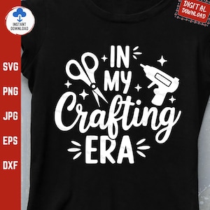 May include: A black t-shirt with white text that reads "In My Crafting Era". The text is surrounded by a pair of scissors, a hot glue gun, and stars.