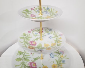 May include: A three-tiered serving stand with a gold-colored handle and support. Each white plate is decorated with a floral pattern in pink, yellow, blue, and green. The stand is designed for displaying desserts or appetizers.