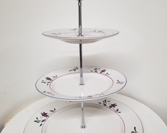 May include: Three-tiered white ceramic serving stand with a chrome metal frame and handle. Each tier features a floral design with red, blue, and green accents.