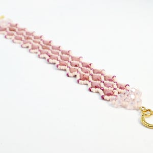 May include: A beaded bracelet featuring a geometric pattern of pink and white beads. The bracelet has a gold-colored clasp and a cluster of clear beads at the end. The bracelet is approximately 7 inches long.