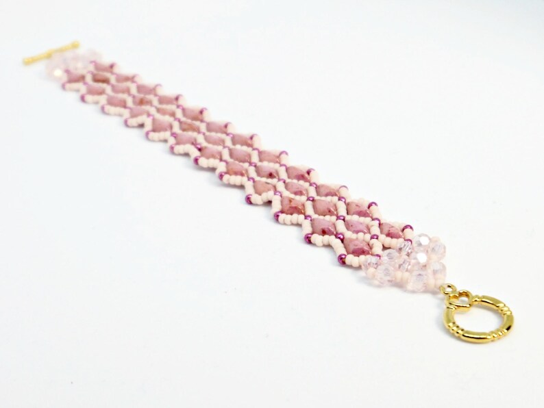 May include: A beaded bracelet featuring a geometric pattern of pink and white beads. The bracelet has a gold-colored clasp and a cluster of clear beads at the end. The bracelet is approximately 7 inches long.