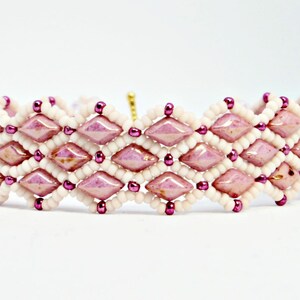 May include: A beaded bracelet featuring a geometric design. The bracelet is composed of alternating rows of light pink diamond-shaped beads and white seed beads, with small purple beads accenting the design. The bracelet is on a white background.