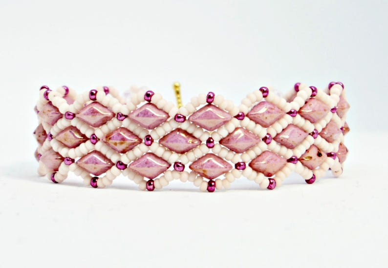 May include: A beaded bracelet featuring a geometric design. The bracelet is composed of alternating rows of light pink diamond-shaped beads and white seed beads, with small purple beads accenting the design. The bracelet is on a white background.