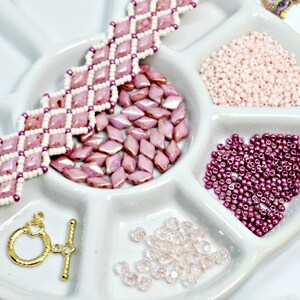 May include: A white ceramic divided tray holds various jewelry-making supplies. A completed pink and white beaded bracelet sits next to loose pink diamond-shaped beads, small pink beads, and a gold-colored clasp. Additional clear and dark pink beads are also present.