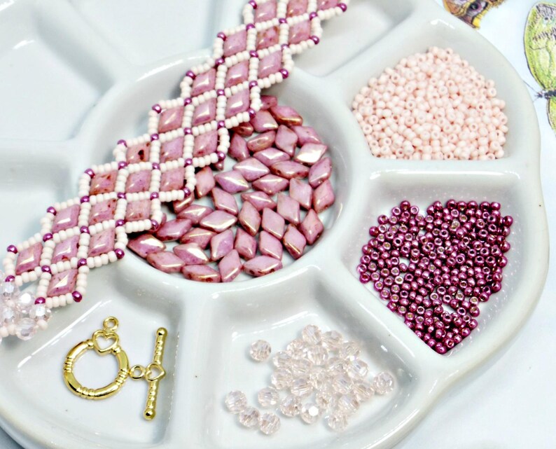 May include: A white ceramic divided tray holds various jewelry-making supplies. A completed pink and white beaded bracelet sits next to loose pink diamond-shaped beads, small pink beads, and a gold-colored clasp. Additional clear and dark pink beads are also present.