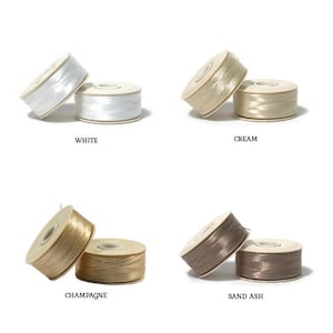 May include: Four spools of decorative ribbon in white, cream, champagne, and sand ash. Each spool is labelled with its colour name. The ribbon is tightly wound on cardboard spools, suitable for crafting and gift wrapping.