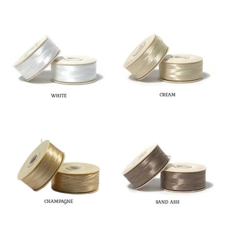 May include: Four spools of decorative ribbon in white, cream, champagne, and sand ash. Each spool is labelled with its colour name. The ribbon is tightly wound on cardboard spools, suitable for crafting and gift wrapping.