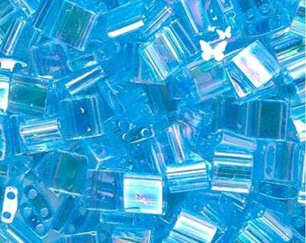 Puede incluir: Una colección de cuentas de vidrio rectangulares de color azul brillante con dos agujeros y un acabado iridiscente. Cada cuenta presenta una superficie translúcida que capta la luz y refleja sutiles tonos arcoíris. Un pequeño gráfico de mariposa blanca está superpuesto.