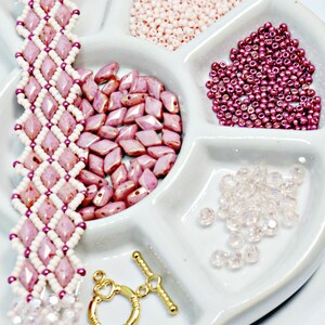 May include: A handmade bracelet with a diamond pattern in pink and white beads. The image also shows various beads in pink and clear, along with a gold-colored clasp, all arranged in a white tray. Jewelry making supplies.