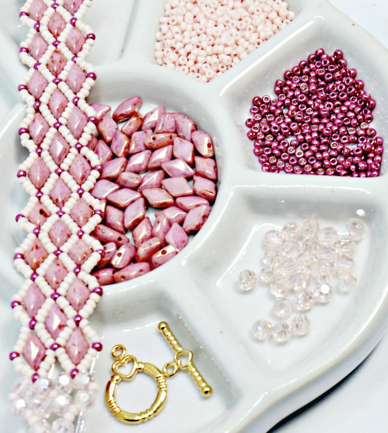 May include: A handmade bracelet with a diamond pattern in pink and white beads. The image also shows various beads in pink and clear, along with a gold-colored clasp, all arranged in a white tray. Jewelry making supplies.