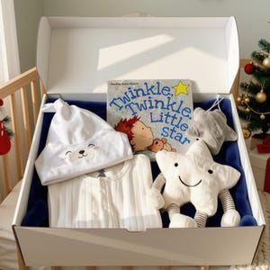 May include: A white gift box containing a white baby hat with a bear face, a white ribbed onesie, a Twinkle Twinkle Little Star book, a plush white star toy with striped arms and legs, and a small grey plush star, all set on a dark blue fabric.