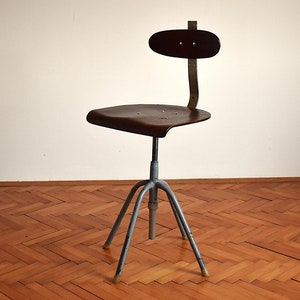 Puede incluir: Una silla giratoria industrial vintage con asiento y respaldo de madera marr&oacute;n oscuro. La silla tiene una base de metal con cuatro patas y una palanca de ajuste de altura.