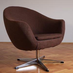 May include: A brown, mid-century modern swivel chair with a curved back and seat. The chair has a textured fabric and sits on a chrome, star-shaped base. The chair is positioned on a wooden floor.
