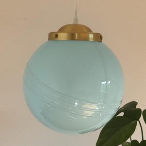 May include: A light blue glass globe pendant light with a gold metal fixture. The light is hanging from a white cord.