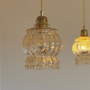May include: Two clear glass pendant lights with a ribbed texture hang from a white ceiling. The larger light has a wider, more rounded shape, while the smaller light is more spherical. Both lights have a brass finish.