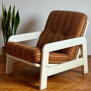May include: A white armchair with a brown and orange striped fabric upholstery. The chair has a simple design with a wide, white frame and a comfortable-looking seat.