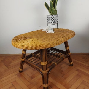 May include: An oval-shaped coffee table constructed from light brown woven wicker with a dark brown bamboo frame. The table features four legs connected by a lower slatted shelf, with light-colored rattan bindings securing the joints throughout.