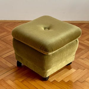 Könnte beinhalten: Ein quadratischer Hocker, bezogen mit olivgrünem Samtstoff. Die Oberseite ist mit einem mittigen Knopf versehen. Das Möbelstück steht auf vier kleinen, dunkel gefärbten Füßen, die eine niedrige Basis für diesen kompakten Gegenstand bilden.