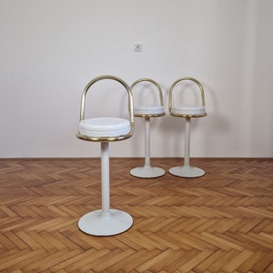 May include: Three white and gold bar stools with a round seat and a curved back. The stools are on a wood floor.
