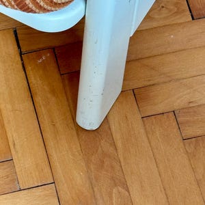 May include: Close-up of a white wooden chair leg with a worn finish. The chair leg is resting on a light brown wood floor.