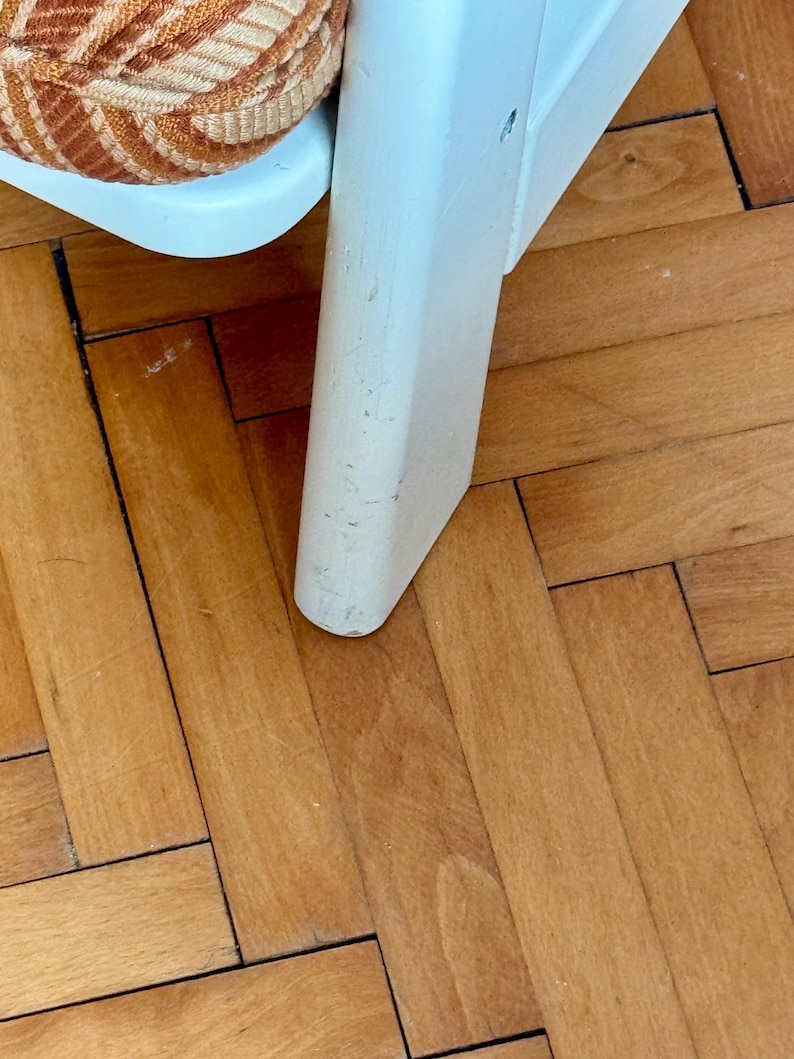 May include: Close-up of a white wooden chair leg with a worn finish. The chair leg is resting on a light brown wood floor.