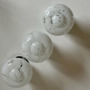 May include: Three white glass cabinet knobs with a speckled pattern. The knobs are round with a slightly domed center and a silver base. The speckled pattern is a mix of white and gray, creating a unique design. The knobs are likely for furniture or cabinetry.