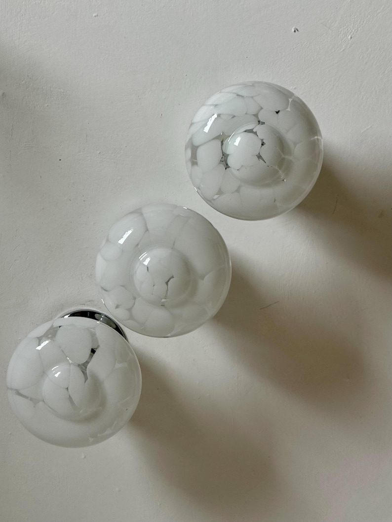 May include: Three white glass cabinet knobs with a speckled pattern. The knobs are round with a slightly domed center and a silver base. The speckled pattern is a mix of white and gray, creating a unique design. The knobs are likely for furniture or cabinetry.